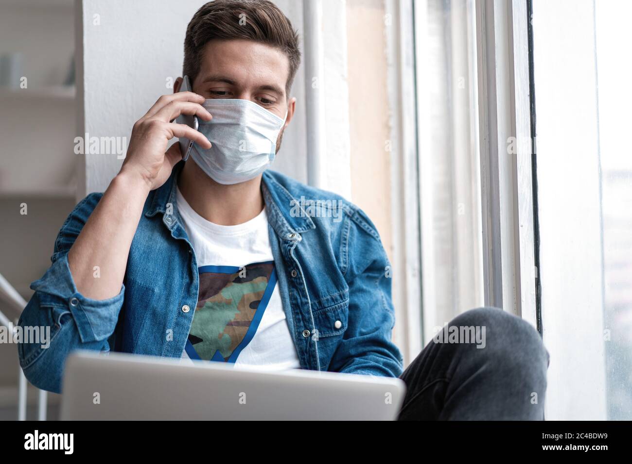 Junger glücklicher Mann sitzt in Schutzmaske am Telefon reden. Stockfoto