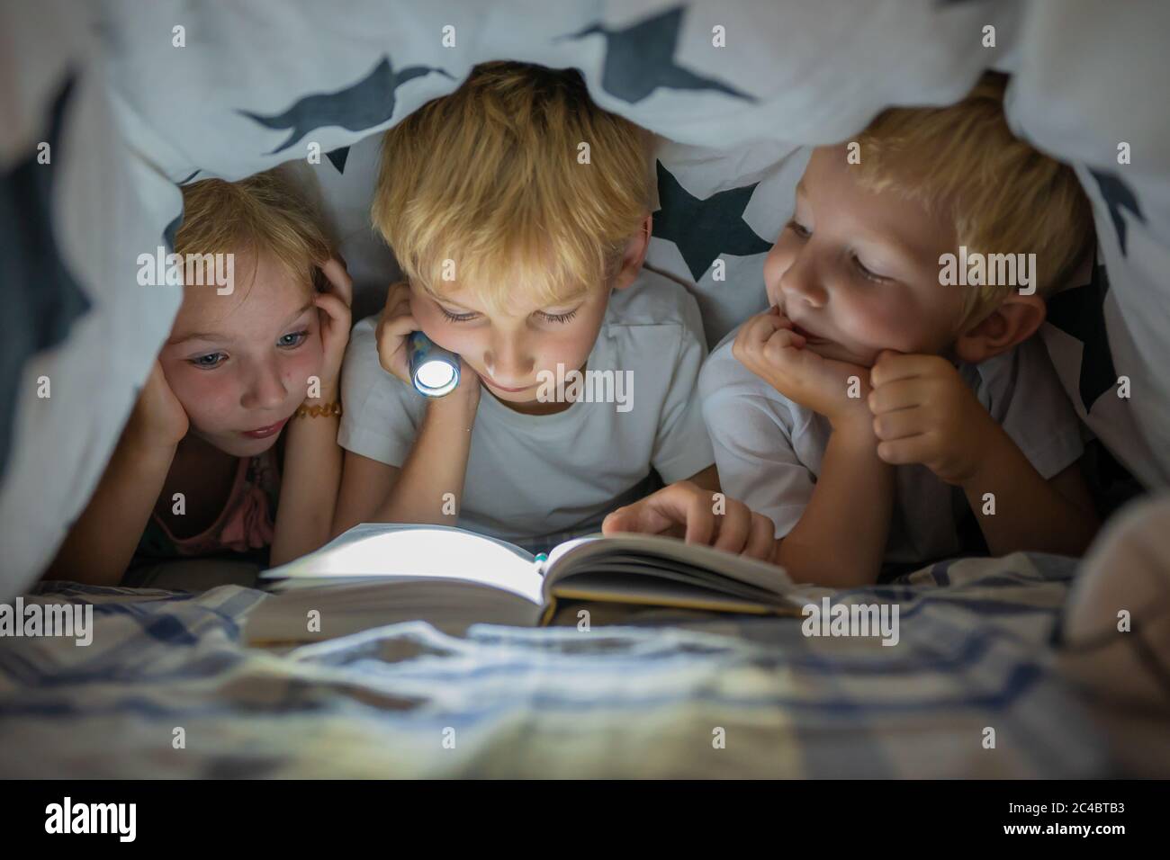Drei kleine Kinder lesen nachts mit einer Taschenlampe unter den Deckblättern ein Buch. Stockfoto
