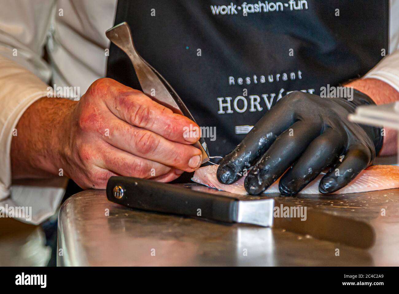 Der Michelin-Sterne-Koch Sebastian Frank entfernt mit einer Fischgrätenzange Gräten aus einem blassen Fischfilet in der Küche des Hotels und Restaurants Kronenschlösschen in Hattenheim, Eltville am Rhein, Hessen, Deutschland. Er hält das Filet mit seiner linken Hand in einem schwarzen Handschuh. Seine schwarze Schürze trägt die Aufschrift „Restaurant Horváth“ und „work·fashion·fun“. Ein Messer liegt vor ihm auf der rostfreien Stahl-Arbeitsfläche. Am Abend des 28. Februar 2017 kocht Frank ein Galadinner auf dem 21. Rheingau Gourmet & Wein Festival. In Österreich geboren, arbeitete er unter anderem im Steirereck in Wien. Stockfoto