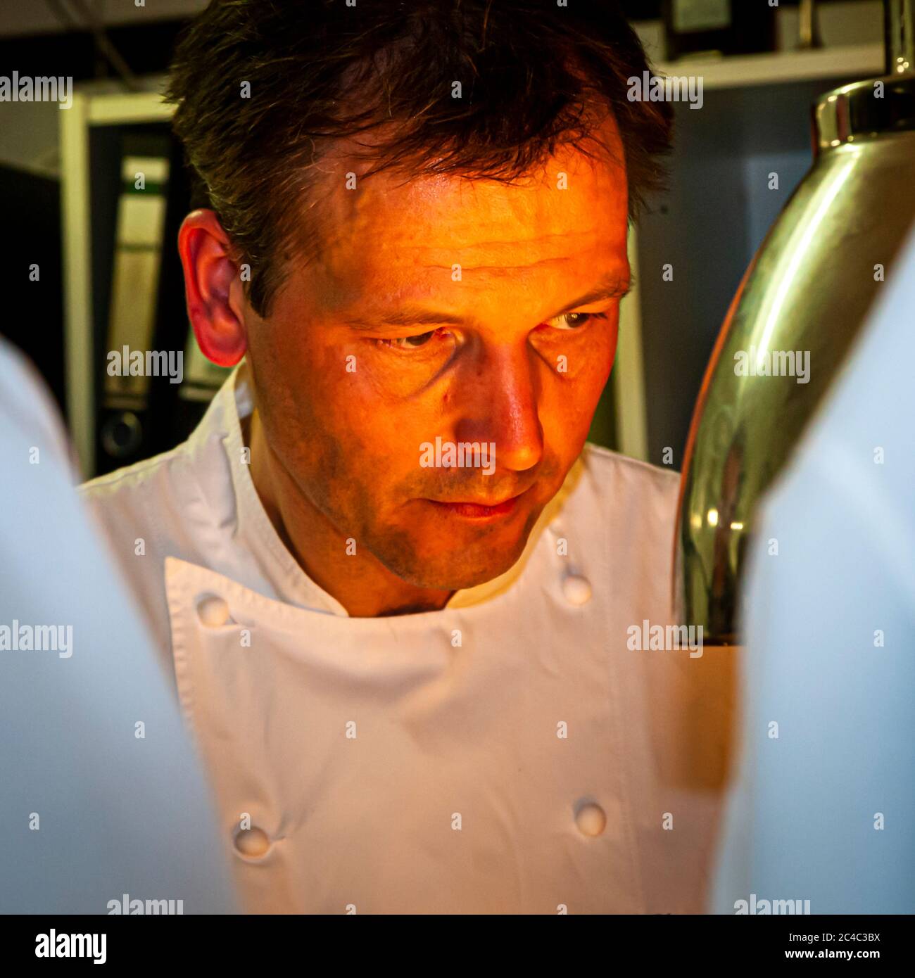 Der mit einem Michelin-Stern ausgezeichnete Koch Thomas Kellermann schaut konzentriert nach unten, sein Gesicht im orangefarbenen Licht einer Wärmelampe am Pass in der Küche des Hotels und Restaurants Kronenschlösschen in Hattenheim, Eltville am Rhein, Hessen, Deutschland. Er trägt eine weiße Kochjacke. Der chromfarbene Schirm der Wärmelampe reicht rechts ins Bild. Thomas Kellermann ist Küchenchef des Restaurants Kastell im Schloss Burg Wernberg in der Oberpfalz, Bayern, wo er seit 2011 zwei Michelin-Sterne hält. Zuvor kochte er unter anderem von 1994 bis 2000 im Tantris in München und war na Stockfoto