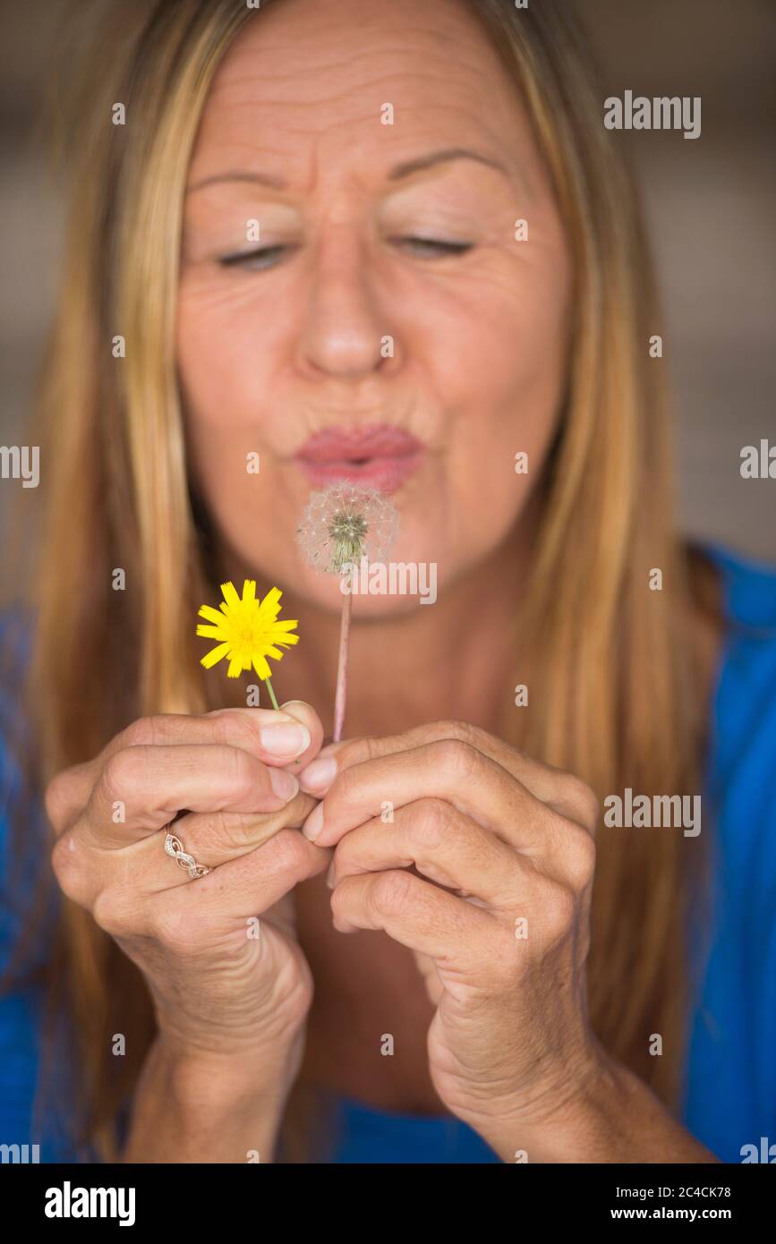 Portrait von attraktiven reifen Frau in verschwommenem Hintergrund weht Löwenzahn Blume in der Hand, glücklich, freundlich. Stockfoto
