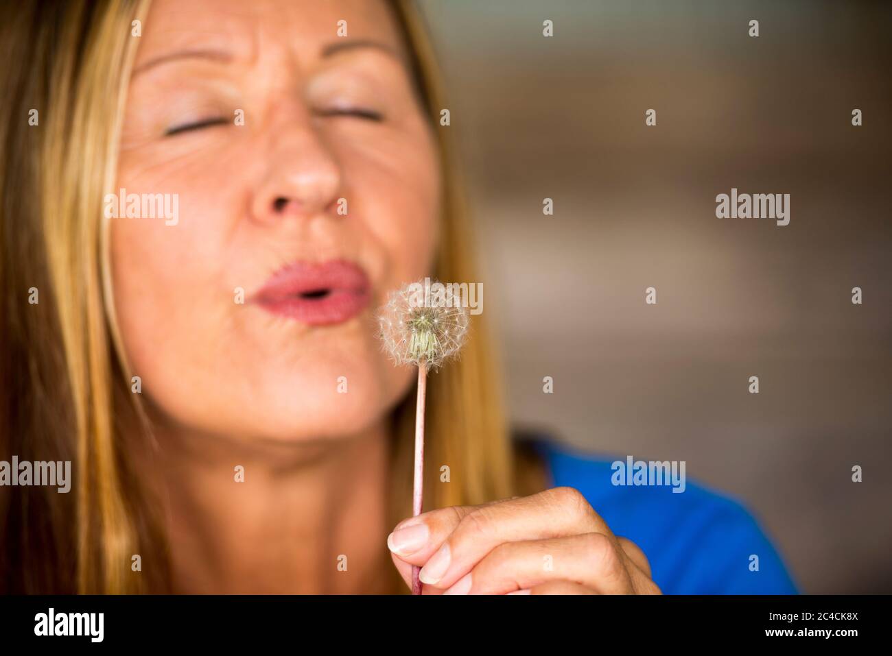 Portrait von attraktiven reifen Frau in verschwommenem Hintergrund weht Löwenzahn Blume in der Hand, glücklich, freundlich, geschlossenen Augen, Copy-Raum. Stockfoto