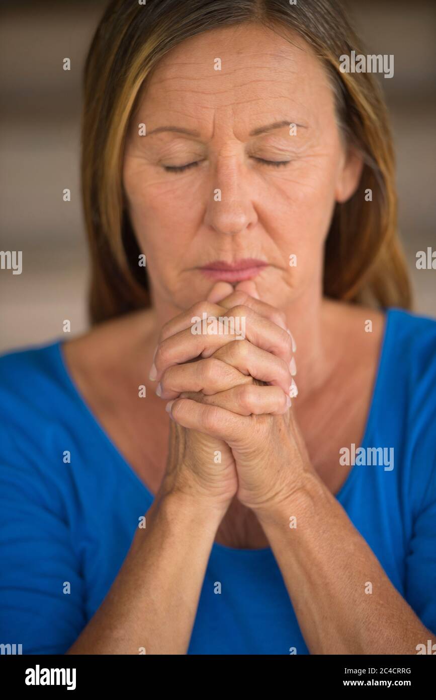 Portrtait Attraktive religiöse reife Frau beten mit Fokus auf gefaltete Hände, nachdenklich, meditierend, geschlossene Augen, verschwommener Hintergrund. Stockfoto