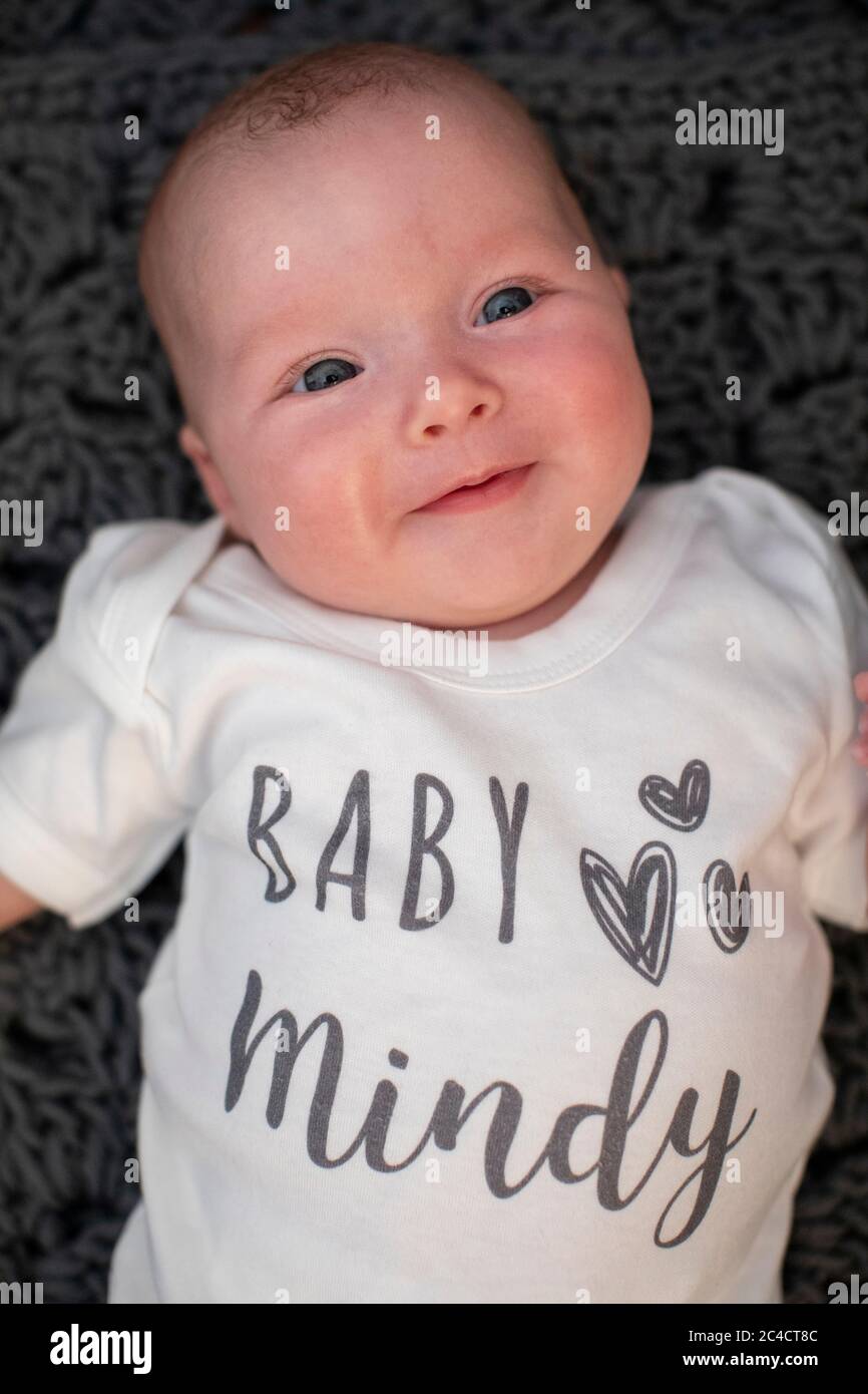 Ein neugeborenes glückliches Baby Mädchen, das auf einem dunklen Teppich mit einer weißen Babyweste mit den Worten Baby Mindy liegt. Foto von Sam Mellish Stockfoto
