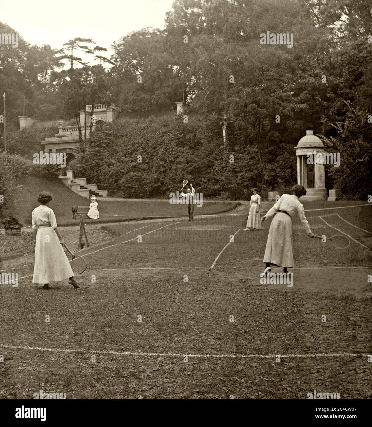 Ein vornehmendes Tennisspiel in Großbritannien um 1900 – ein Mann und drei Damen spielen in einem Landhaus/Herrschaftsgarten auf einem sehr unebenen Rasen. Der Sport sieht hier wie ein Zeitvertreib der Oberschicht aus. Stockfoto
