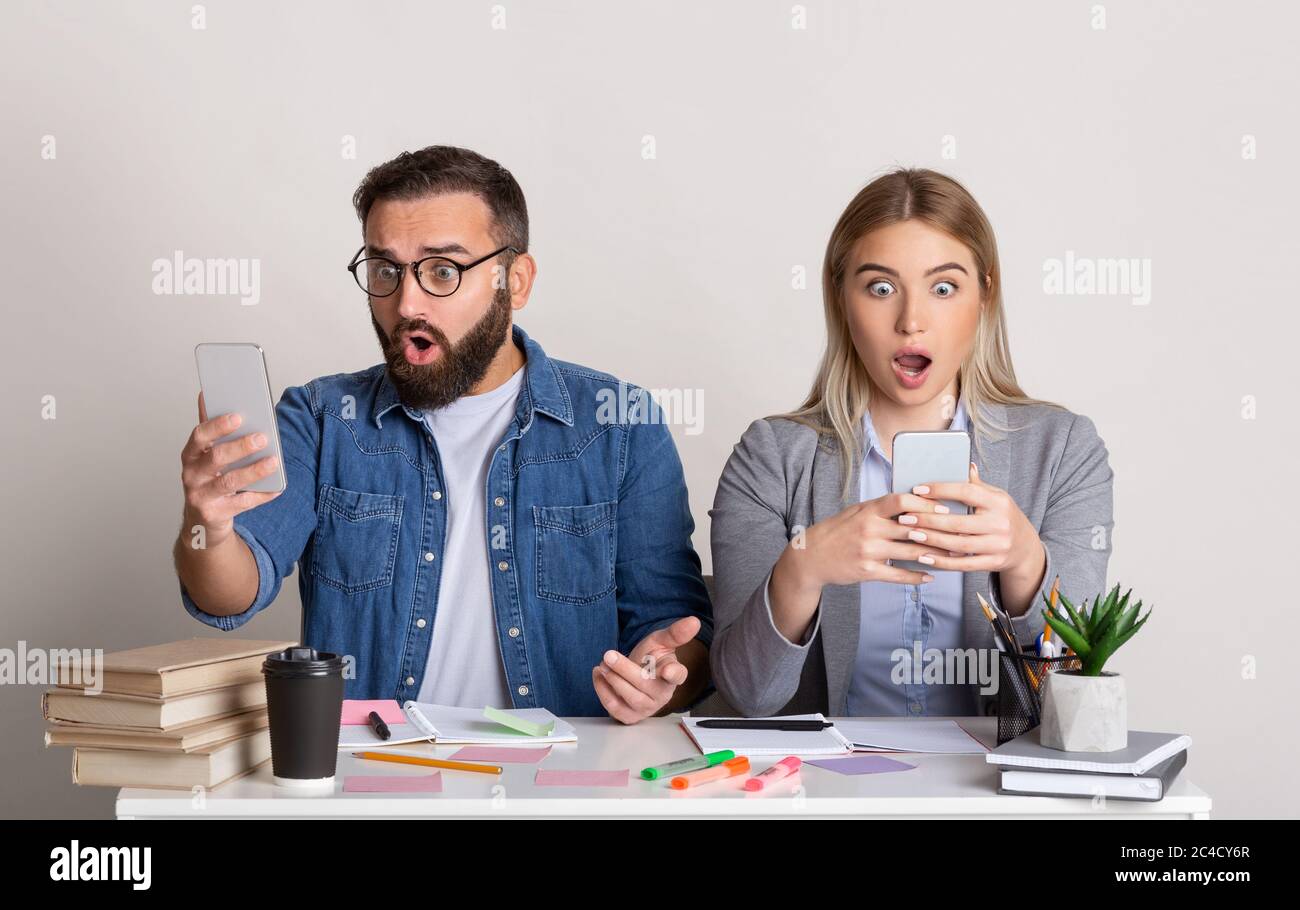 Schockierende Nachrichten am Arbeitsplatz. Kollegen Kerl und Mädchen sitzen in der Nähe, mit offenem Mund und Blick auf Smartphones Stockfoto