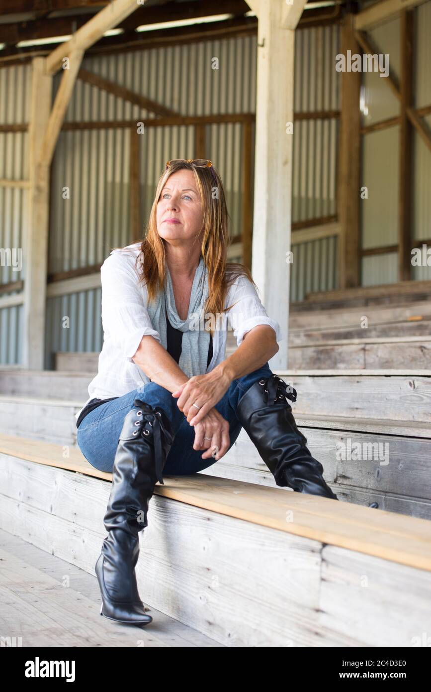 Portrait Attraktive reife Frau, die nachdenklich und besorgt auf der Bank sitzt, Jeans, Bluse und Stiletto-Stiefel trägt. Stockfoto