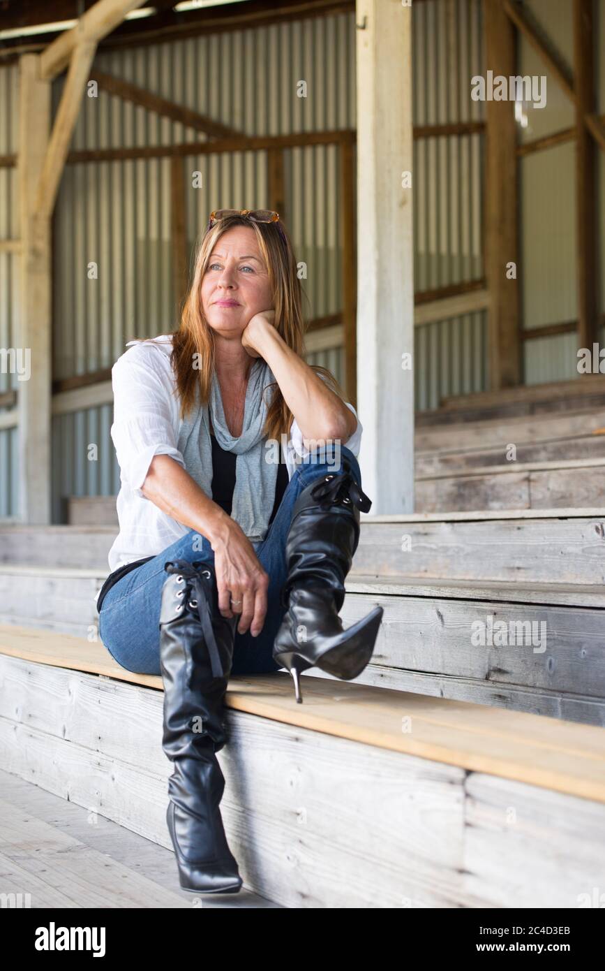 Portrait Attraktive reife Frau entspannt sitzend besorgt, aber nachdenklich auf der Bank, trägt Jeans, Bluse und Stiletto Stiefel. Stockfoto