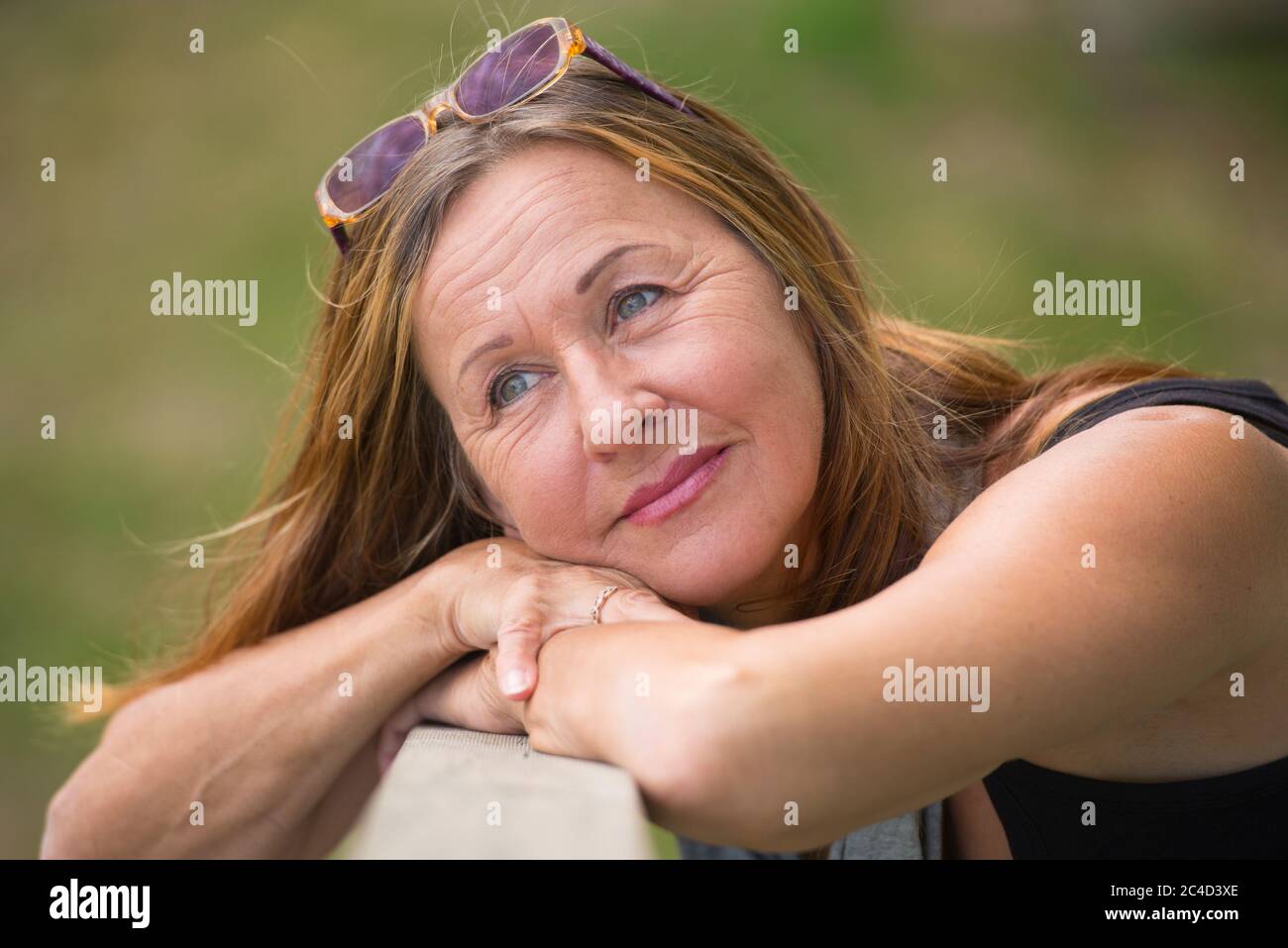 Portrait Attraktive reife Frau posiert entspannt und glücklich lächelnd im Freien mit Sonnenbrille, grün verschwommener Hintergrund und Kopierer Raum. Stockfoto