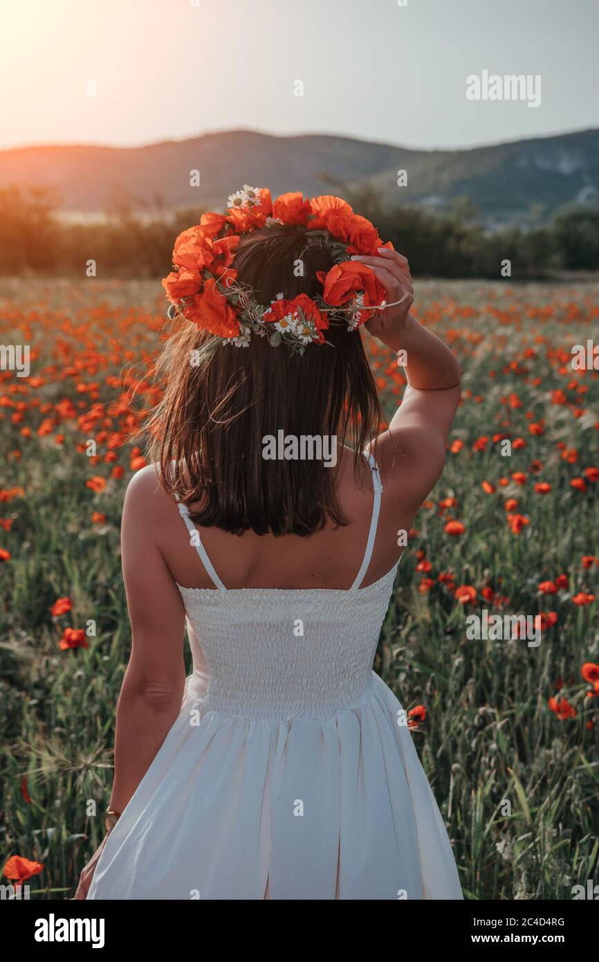 Braut in einem weißen Kleid in einem Kranz aus roten Mohnblumen, warme Sonnenuntergangszeit auf dem Hintergrund des großen roten Mohnfeldes. Speicherplatz kopieren. Das Konzept der Ruhe Stockfoto