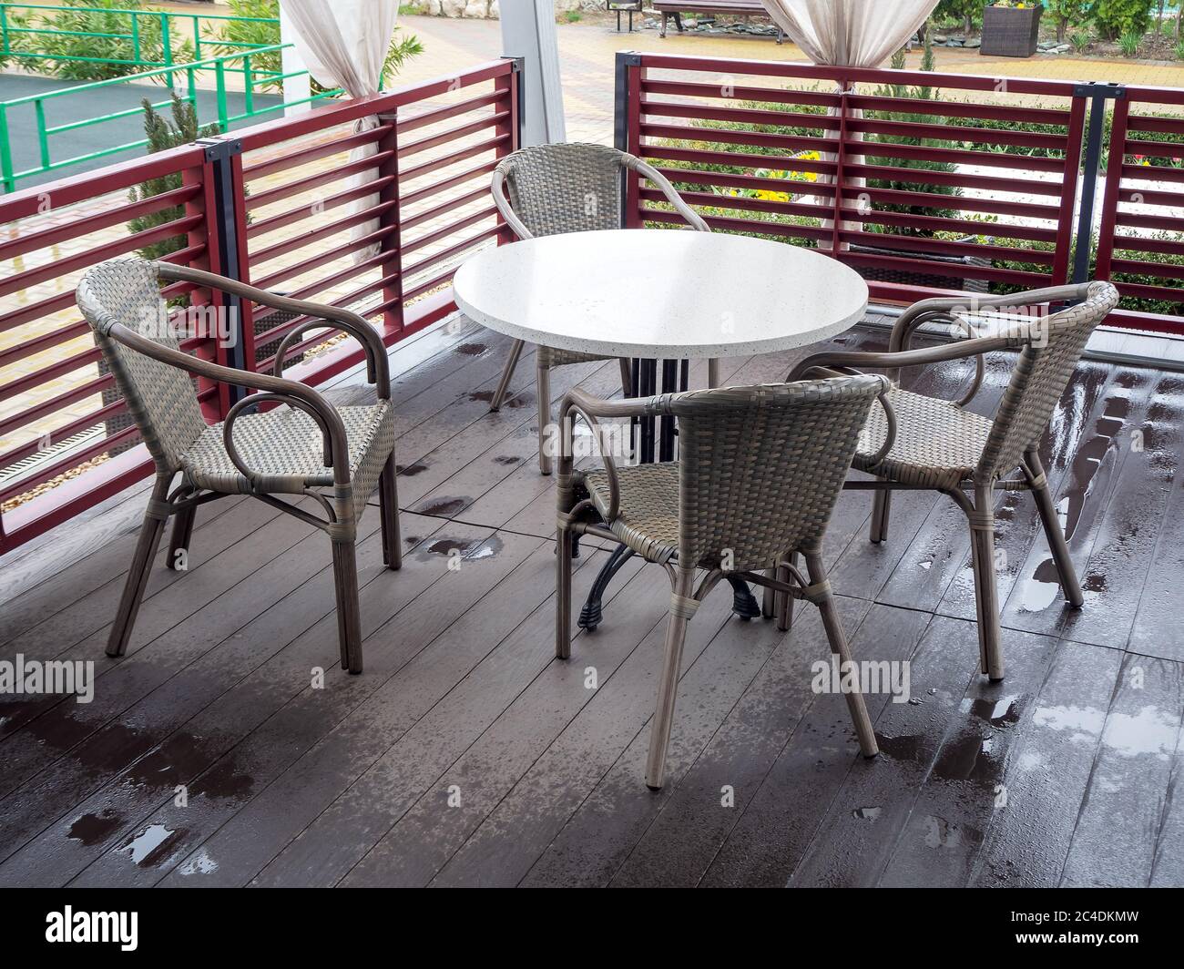 Korbsessel stehen um einen weißen runden Tisch auf einem Holzboden, der nach Regen in einem Café im Freien nass ist Stockfoto