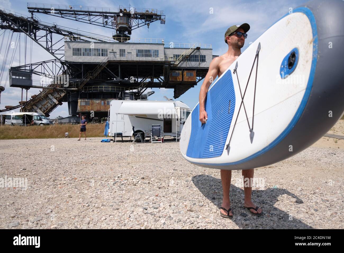 26. Juni 2020, Sachsen-Anhalt, Gräfenhainichen: Ein Urlauber steht in Ferropolis mit einem Standup Paddle Board (SUP) vor stillgestandem Bergbaugerät. Wegen Corona gibt es in diesem Jahr keine großen Festivals. So genanntes Pop-up-Camping soll Urlauber in die Gegend locken. Es gibt 50,000 Stellplätze für Campingfans auf dem Gelände. Foto: Sebastian Willnow/dpa-Zentralbild/dpa Stockfoto