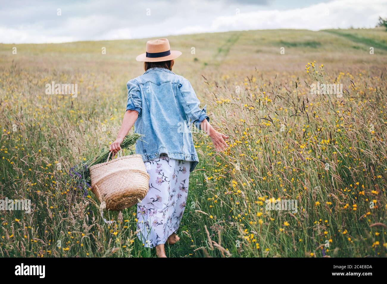 Junge Frau gekleidet Jeans Jacke und leichte Sommerkleid zu Fuß durch die hohe grüne Wiese mit Korb und Wildblumen Bouquet. Mensch und Natur c Stockfoto