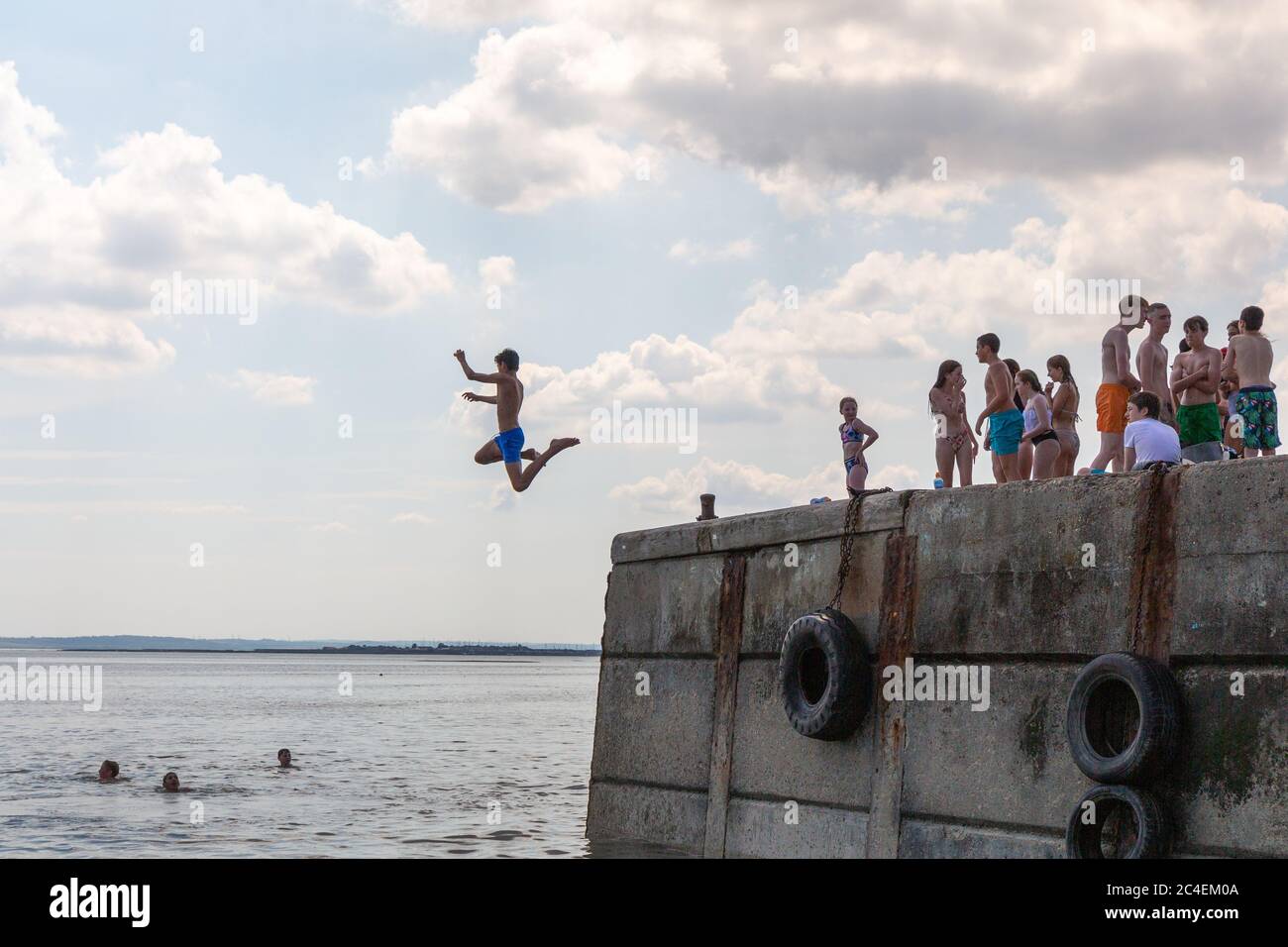 Leigh-on-Sea, Großbritannien. Juni 2020. Junge Leute springen vom Betonpier ins Meer am Bell Wharf Beach, Leigh-on-Sea, Essex. Andere schwimmen oder beobachten vom Dock aus, genießen warmes Wetter und Freizeit an der Küste. Große Gummireifen säumen die Kante des Piers und deuten auf ihre Verwendung als Festmacher hin. Die Szene fängt spontane Freude, Erholung am Meer und Gemeinschaftsgeist ein. Die Leute springen von der Bell Wharf ins kühle Wasser. Leute in Old Leigh, Essex, genießen das warme Wetter und weniger überfüllte Strände. Penelope Barritt/Alamy Live News Stockfoto
