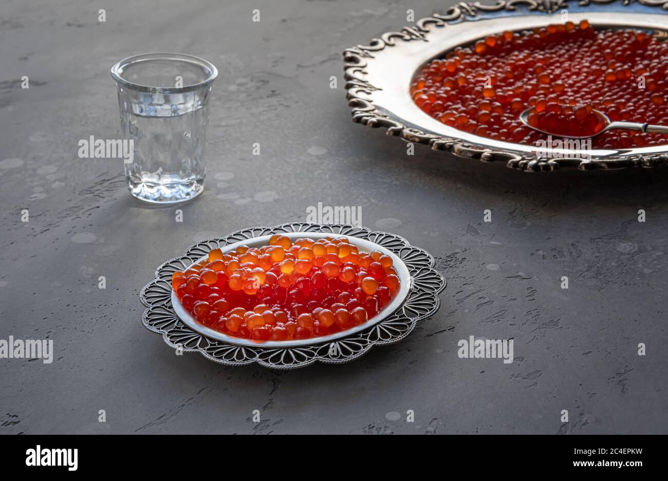 Natürlicher roter Kaviar auf silbernem Teller auf grauem Hintergrund. Wodka Schüsse und Teller mit Kaviar auf unfokussierten Hintergrund. Selektiver Fokus. Gesunde Ernährung concep Stockfoto