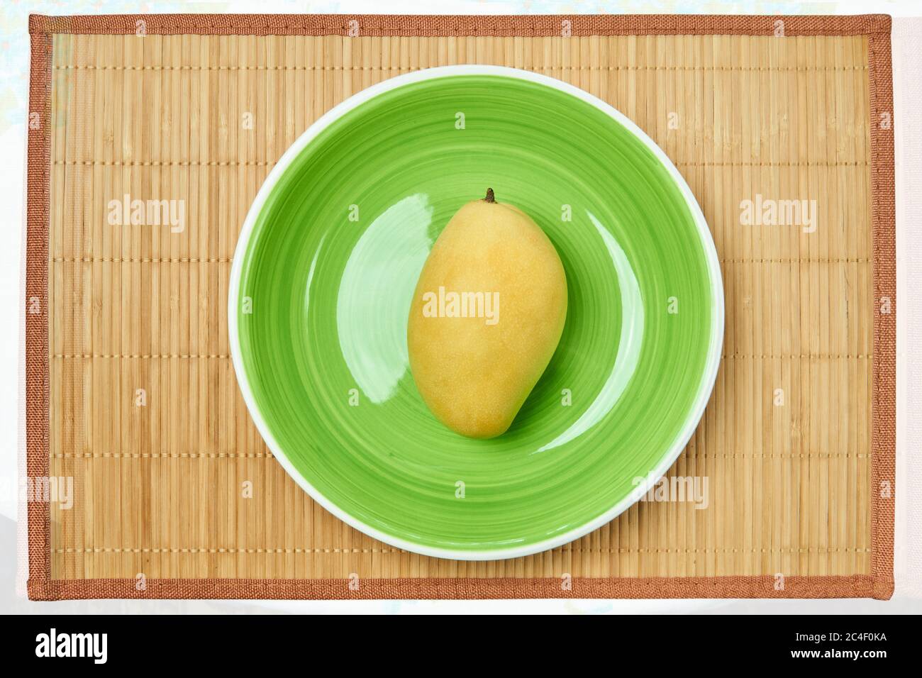 Farbe Stillleben - gelbe Mangofrucht auf einem grünen Teller auf einer Rohrservierung Matte Stockfoto