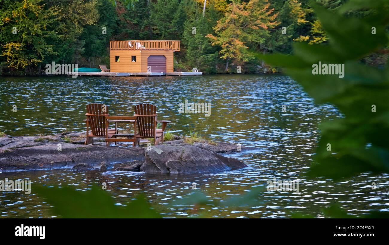 Retirement Living - zwei Muskoka Stühle sitzen auf einem felsigen Ufer mit Blick auf einen ruhigen See mit Bäumen und einem Boot Stockfoto