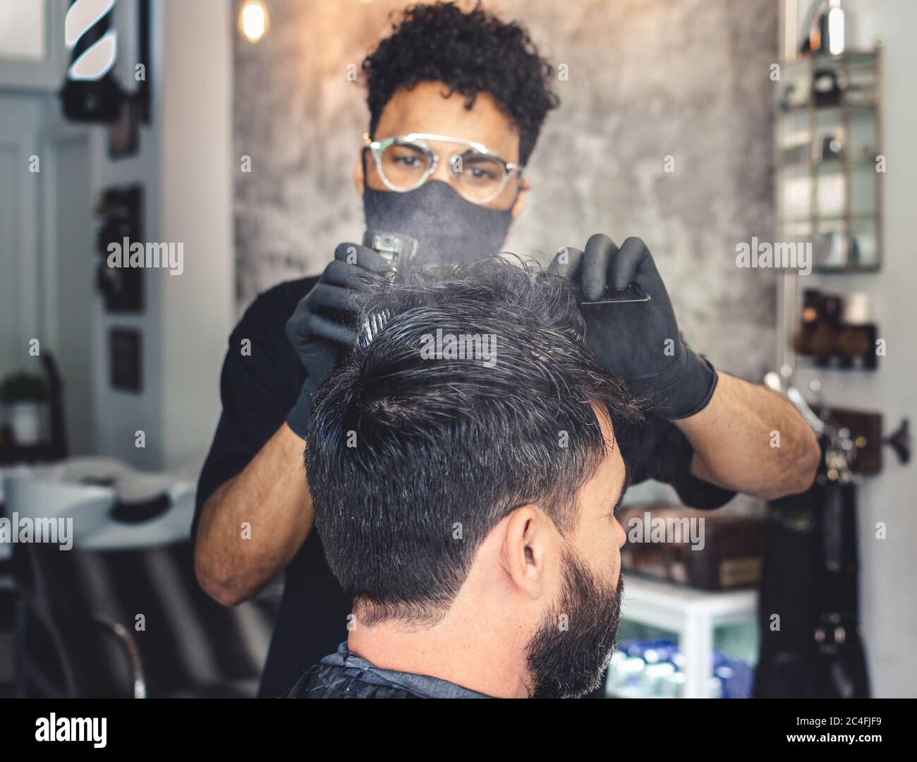 Mulatto Barber mit einer Maske Benetzung eines Kunden Haar mit einem Sprüher aus dem Hintergrund Stockfoto