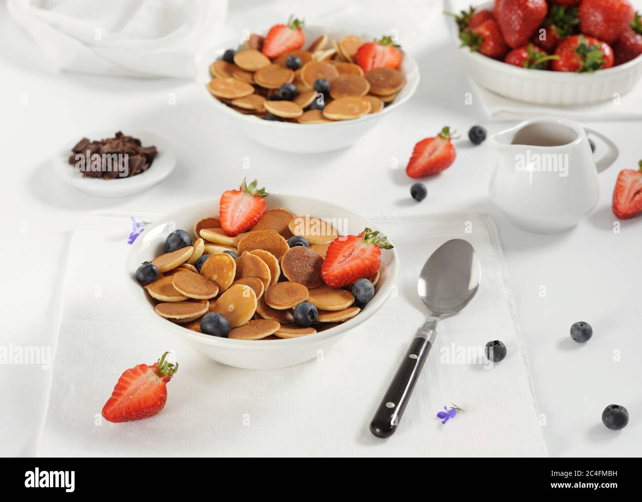 Süße hausgemachte Schale mit winzigen Pfannkuchen mit Schokolade, Honig, Erdbeeren und Heidelbeeren auf weißem Tisch. Nahaufnahme.Horizontal, selektiver Fokus. Stockfoto