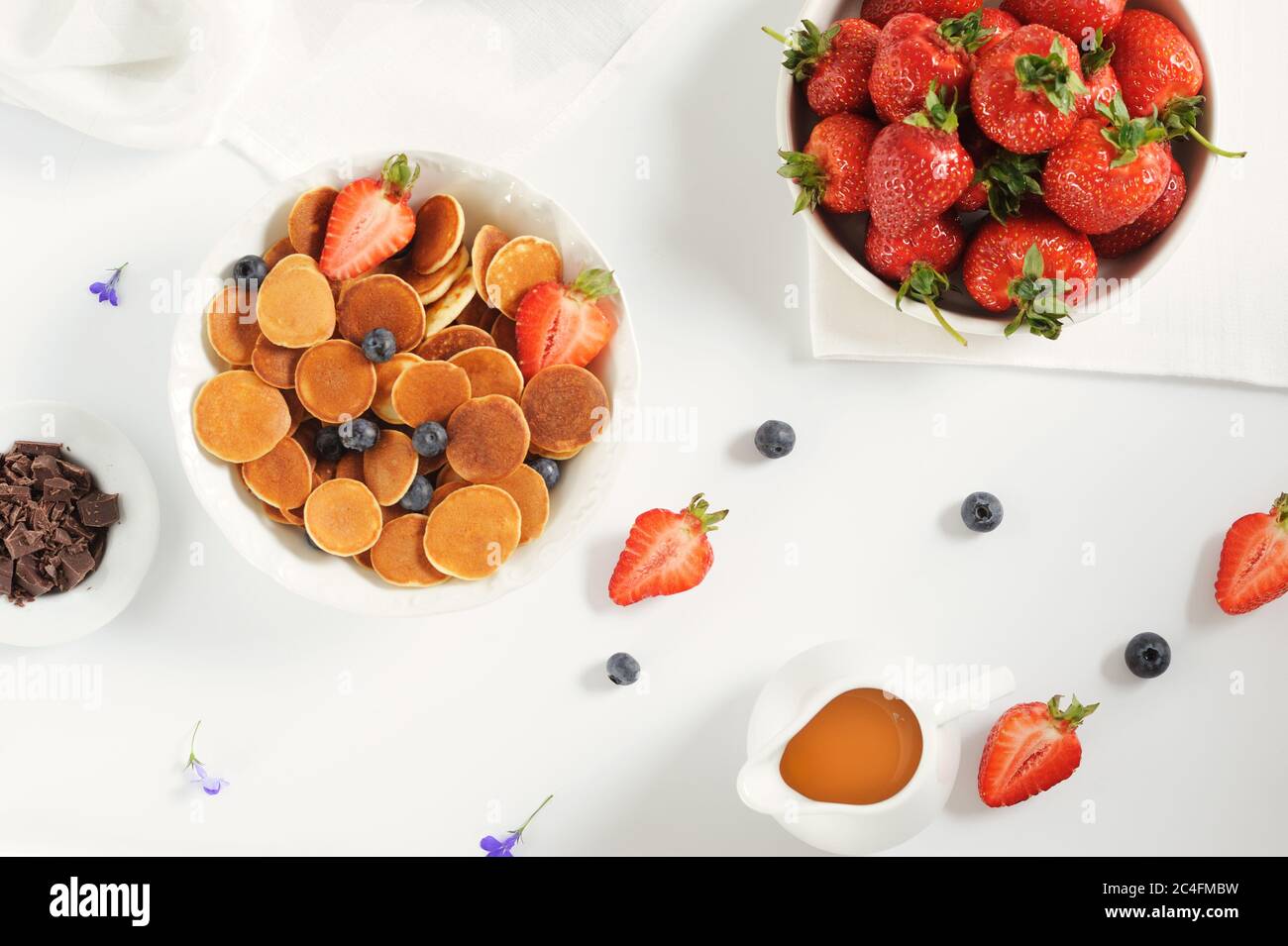 Süße hausgemachte Schale mit winzigen Pfannkuchen mit Schokolade, Honig, Erdbeeren und Heidelbeeren auf weißem Tisch. Flach liegend. Horizontaler, selektiver Fokus. Stockfoto