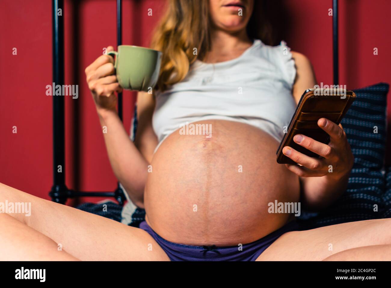 Eine junge schwangere Frau trinkt aus einem Becher und benutzt ihr Smartphone im Bett Stockfoto