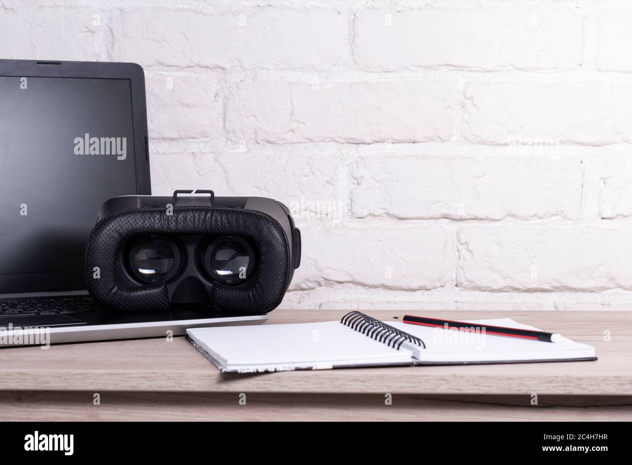 VR-Gerät mit Laptop auf einem Holztisch in der Nähe einer weißen Ziegelwand Stockfoto