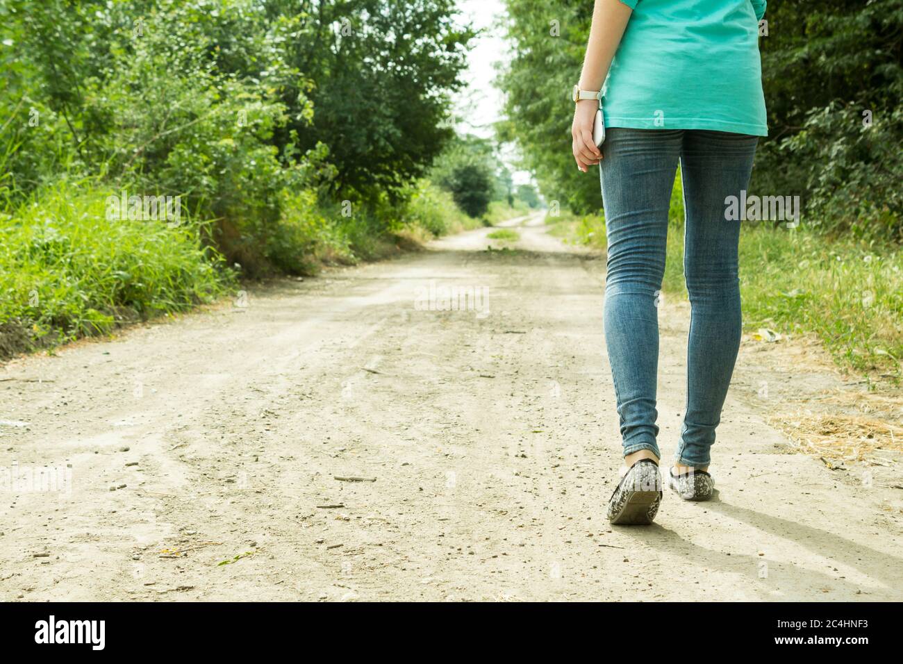 Das Mädchen geht auf einem Feldweg mit einem Telefon in der Hand mit Aussicht in die Ferne durch die Bäume, Rückansicht Stockfoto