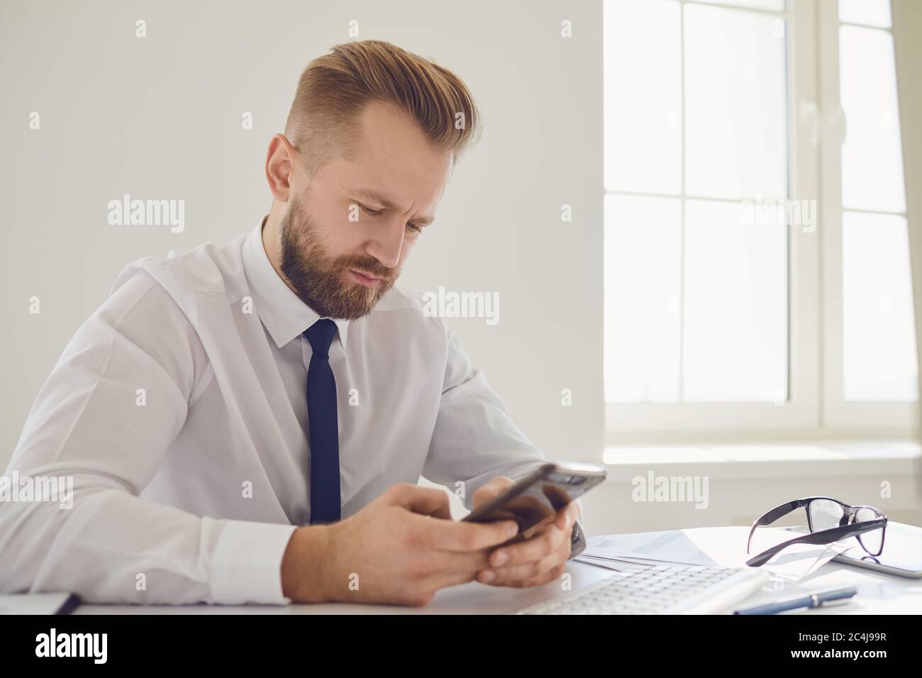 Ein Unternehmer, der mit viel beschäftigt ist, liest die Nachricht am Telefon an einem Tisch mit einem Computer im Büro. Stockfoto