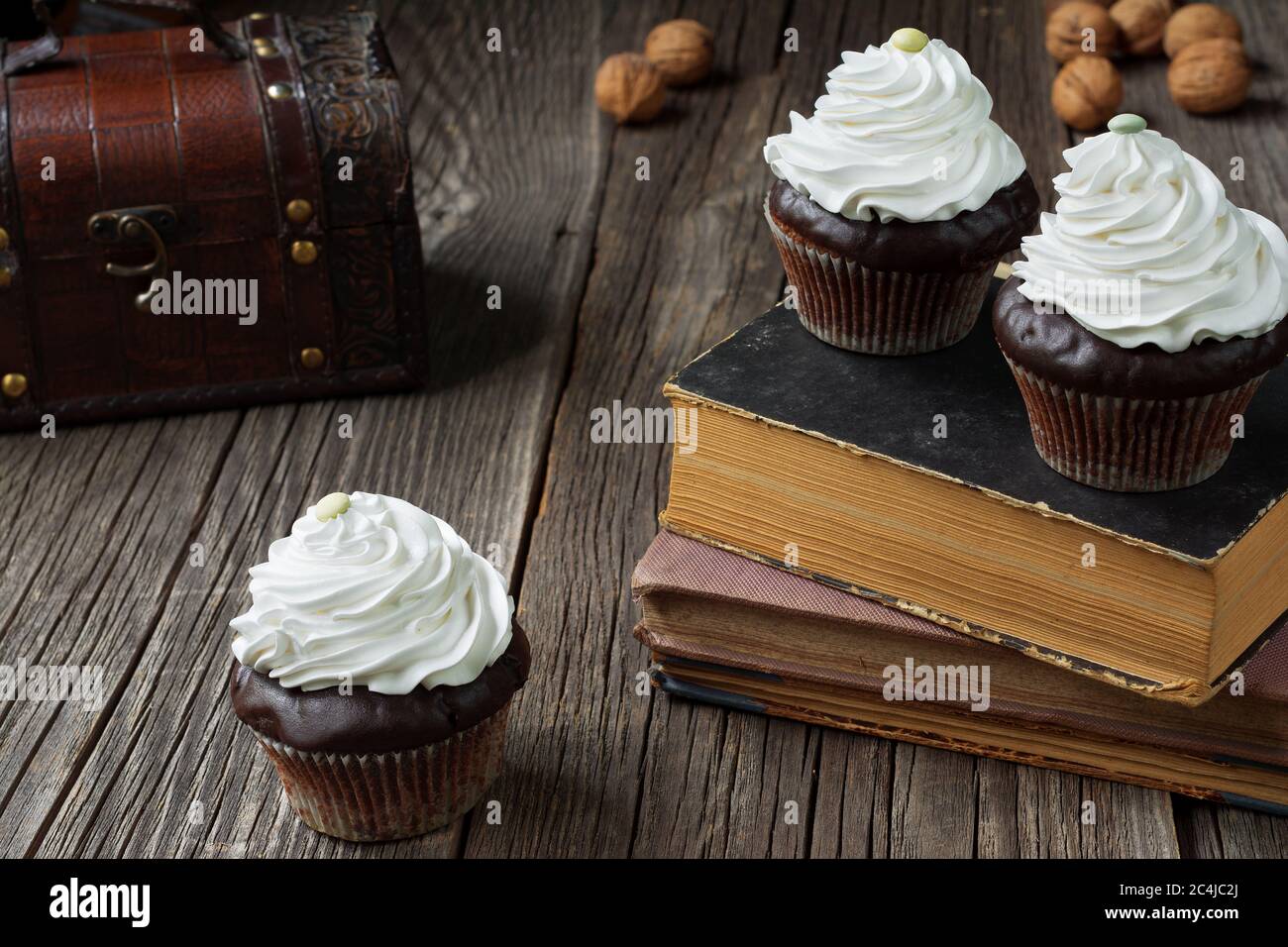 Cupcakes in Pappkisten, mit Schlagsahne überbacken, liegen auf antiken Büchern und verwitterten Holzplanken. Eine Brust und ein paar Walnussschalen... Stockfoto