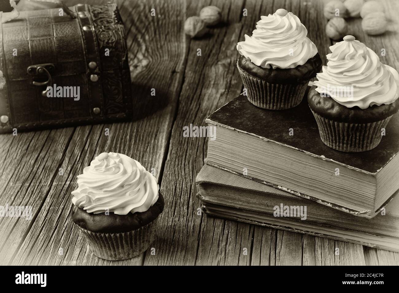 Cupcakes in Pappkisten liegen auf alten Büchern und verwitterten Holzplanken. Eine Brust und ein paar Walnussschalen...EIN Schwarz-Weiß-Fotoeffekt Stockfotografie
