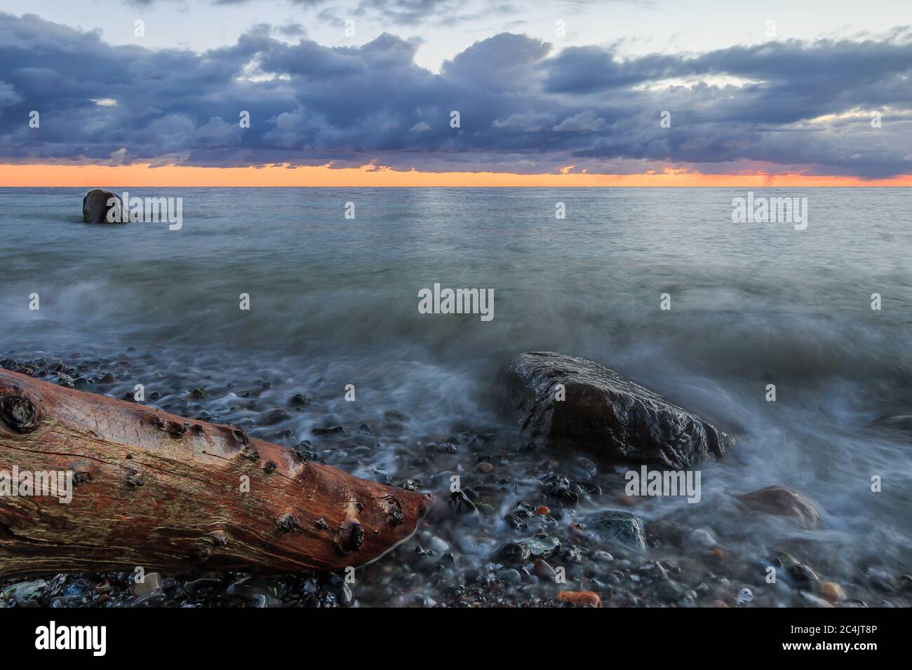 Küstenlinie am Morgen an der Ostsee. Dramatische Wolken bei Sonnenaufgang. Steinige Küste auf der Insel Rügen mit altem Baumstamm und großen Steinen Stockfoto
