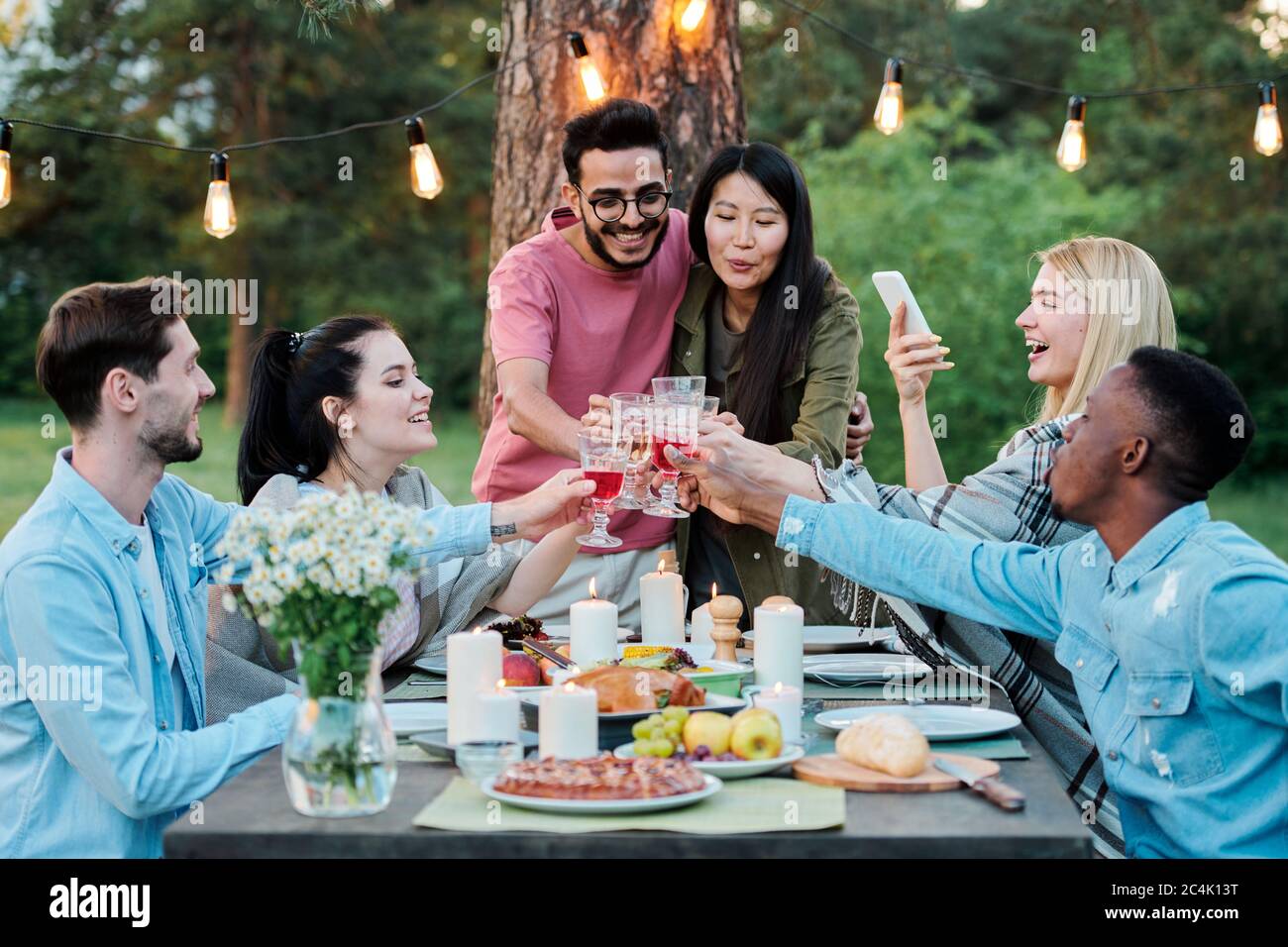 Gruppe interkultureller fröhlicher Freunde versammelt von servierten Tisch unter Baum klirend mit Gläsern mit hausgemachtem Wein Stockfoto