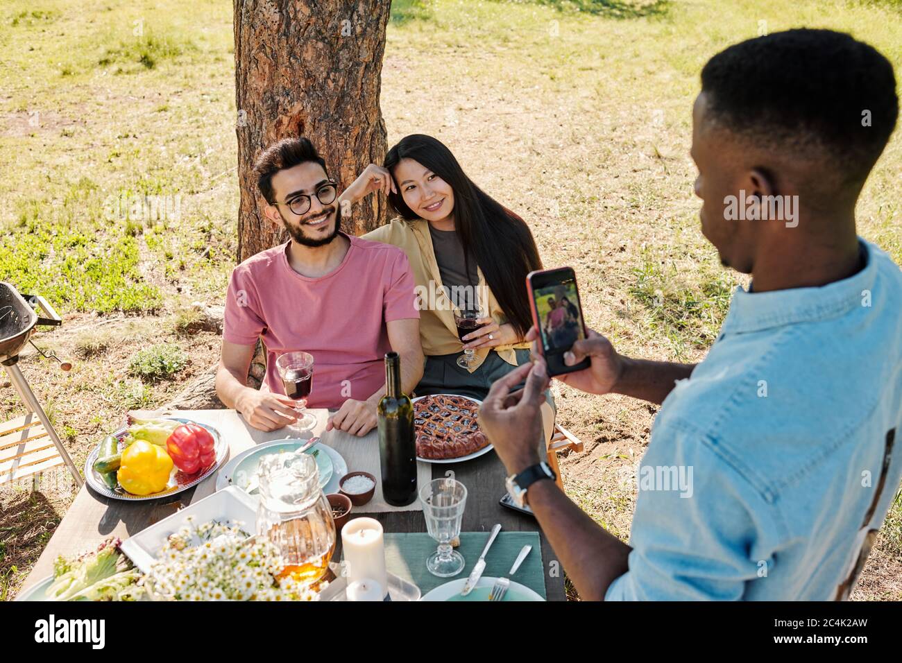 Junger Afrikaner mit Smartphone fotografiert interkulturelles Paar sitzt unter Kiefern vor dem Tisch serviert zum Abendessen Stockfoto