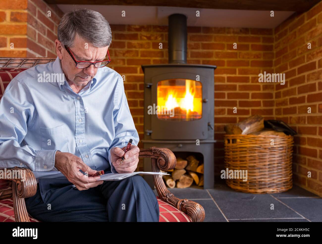 Älterer kaukasischer Erwachsener, der im Sitzen ein Dokument mit einem Füllfederhalter in der Hand überprüft. Er sitzt am Holzofen Stockfoto