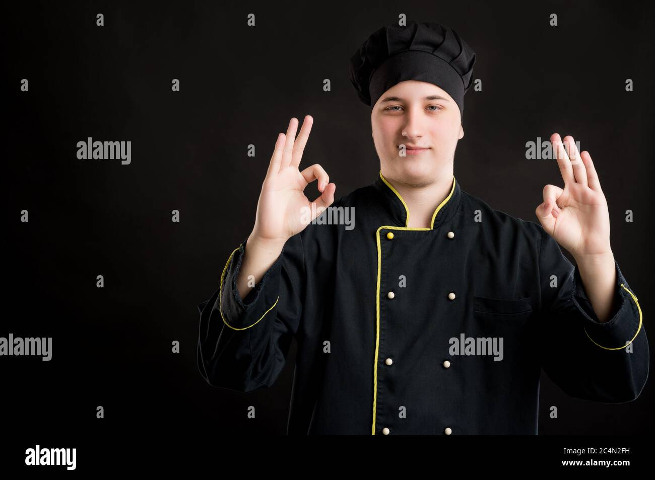 Portrait von jungen Mann in einem schwarzen Koch Anzug gekleidet zeigt Doppel OK-Zeichen posiert auf einem schwarzen isolierten Hintergrund mit Kopie Raum Werbefläche Stockfoto
