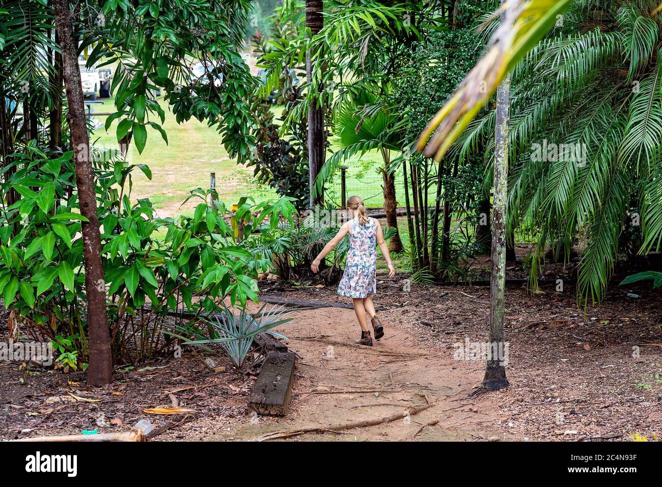 Townsville, Queensland, Australien - Juni 2020: Junge blonde Mädchen in einem bunten Kleid und Stiefel geht einen Feldweg in den Garten ihres Hauses Stockfoto