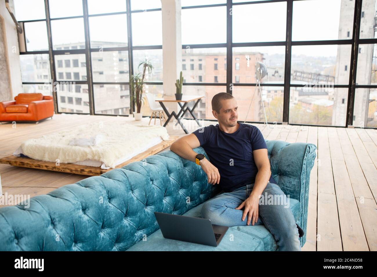 Mann, der auf der Couch mit Laptop sitzt und im Fenster schaut Stockfoto