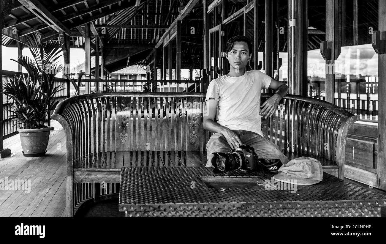 Miri, Sarawak, Malaysia: Ein Mann mit Kamera sitzt auf einer Bank des kunstvoll geschnitzten hölzernen Veranstaltungssaals im 'Coco Cabana Miri' Stockfoto