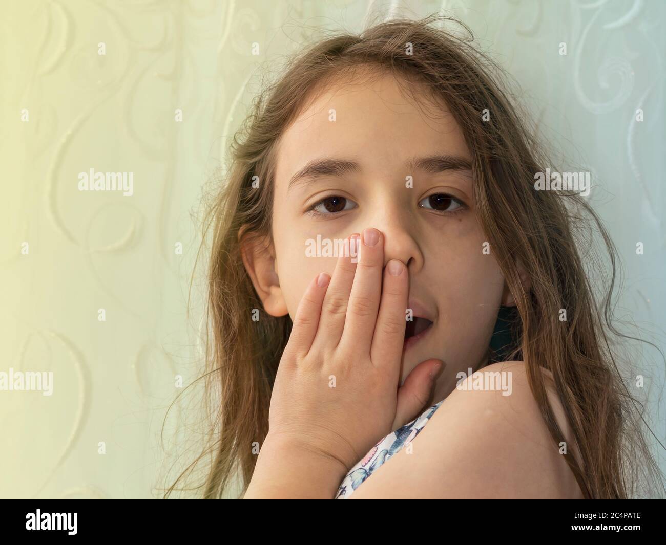 Ein kleines Mädchen schließt ihren eigenen Mund. Erstaunt und überrascht Langhaar Mädchen schließen ihren eigenen Mund mit ihrer eigenen Hand vor dem gelben und blauen Hintergrund Stockfoto