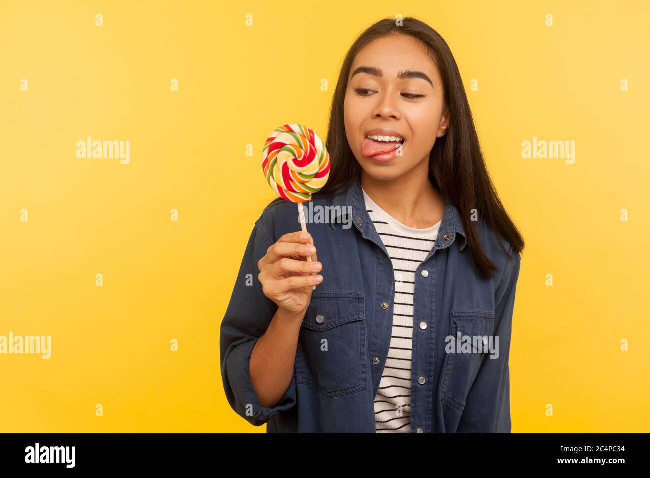 Portrait von glücklich sorglos Mädchen in Denim-Shirt stehen mit Zunge aus, träumen und versucht, Lollipop, rund süß zuckersüßen Regenbogen Süßigkeiten schmecken. ind Stockfoto