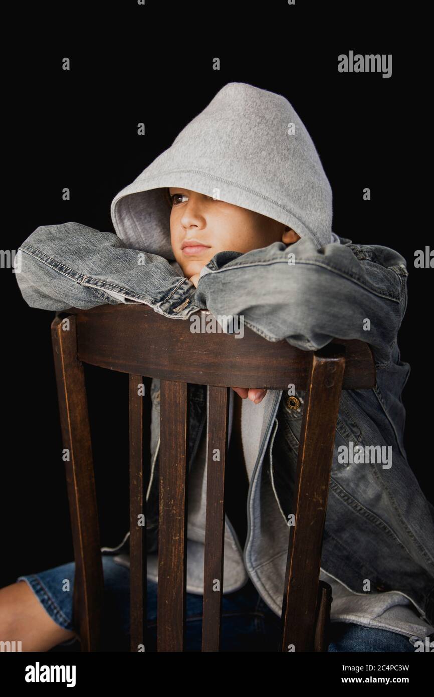 Elf Jahre alter Junge mit Kapuzenpullover und einer jean-Jacke auf einem Holzstuhl vor schwarzem Hintergrund sitzen Stockfoto