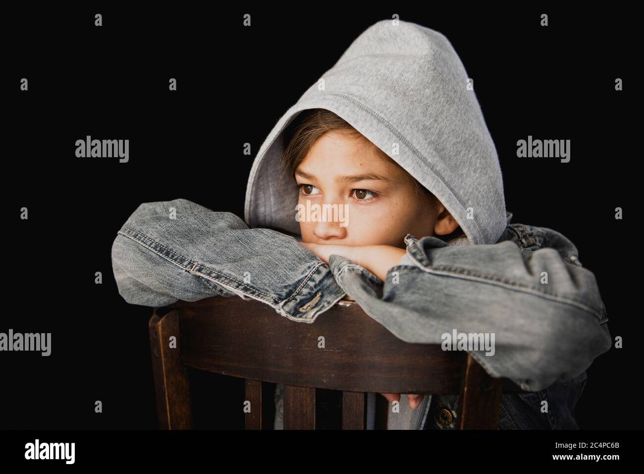 Elf Jahre alter Junge mit Kapuzenpullover und einer jean-Jacke auf einem Holzstuhl vor schwarzem Hintergrund sitzen Stockfoto