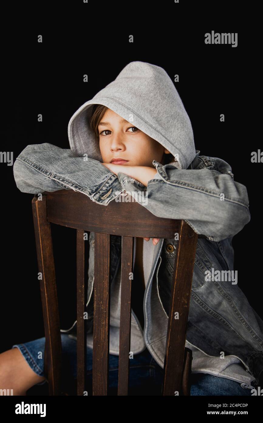 Elf Jahre alter Junge mit Kapuzenpullover und einer jean-Jacke auf einem Holzstuhl vor schwarzem Hintergrund sitzen Stockfoto