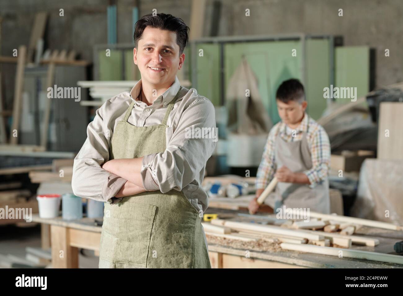 Portrait eines lächelnden, selbstbewussten Tischlers mittleren Alters in Schürze, der mit gekreuzten Armen in der Werkstatt steht, sein Sohn arbeitet mit Holz im Hintergrund Stockfoto