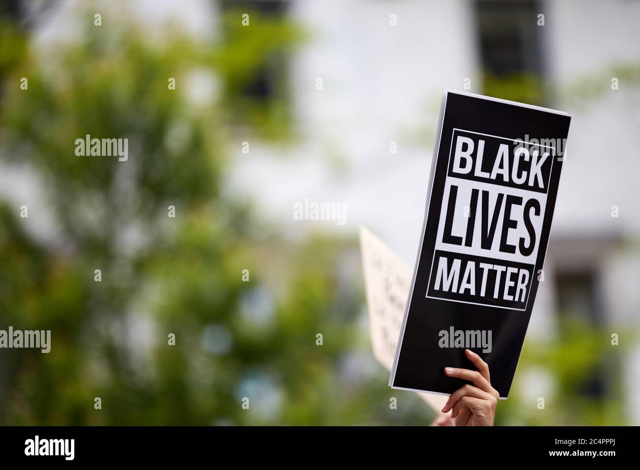 Eine Person, die ein gedrucktes schwarzes Schild "Black Lives Matter" bei einer rassisch orientierten Kundgebung hält. Ein verschwommenes Gebäude und Bäume im Hintergrund. Aktivismus Stockfoto