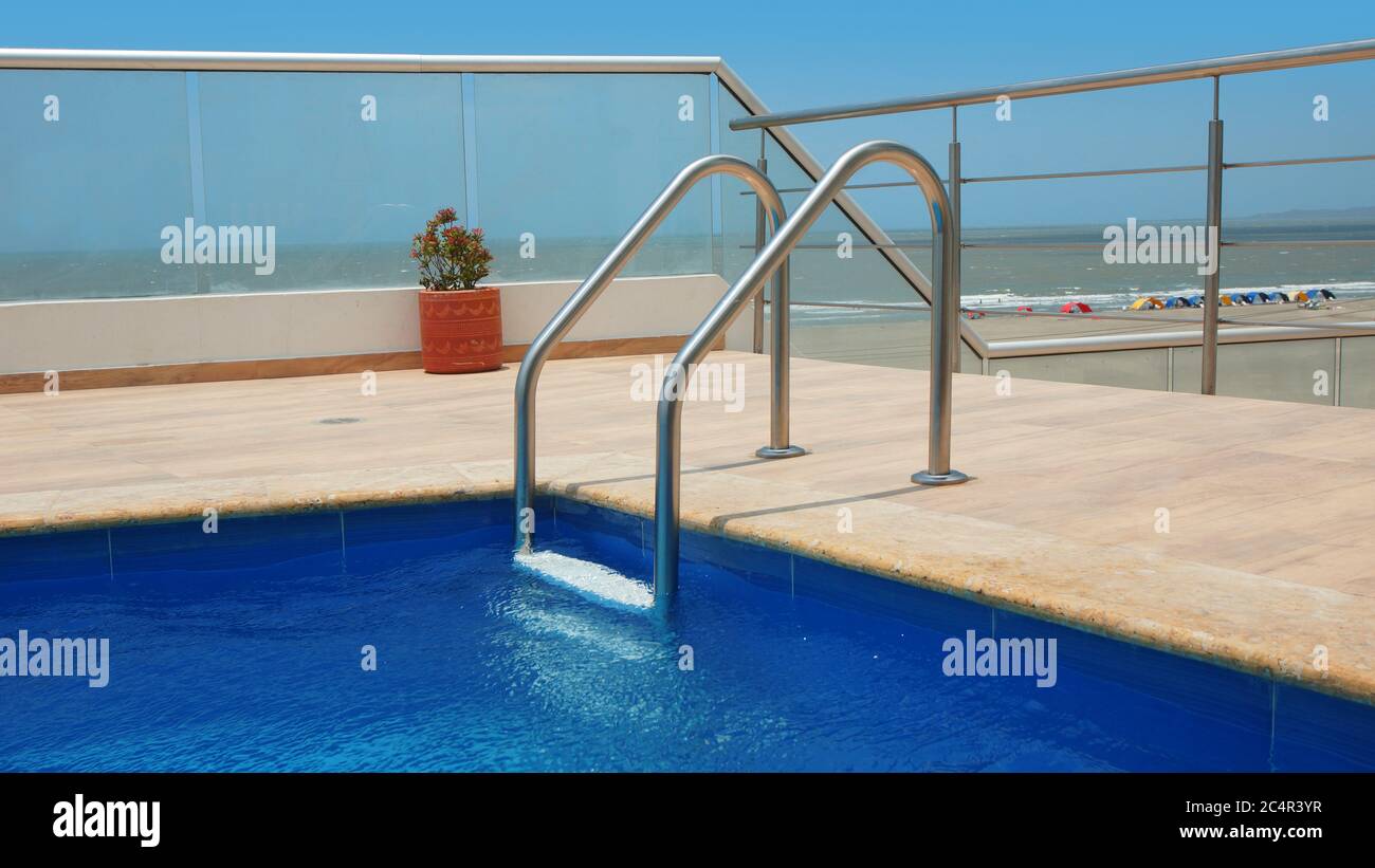Ecke eines Schwimmbades mit Marmorrand, blauen Keramikwänden und Poolleiter mit Strand im Hintergrund Stockfoto