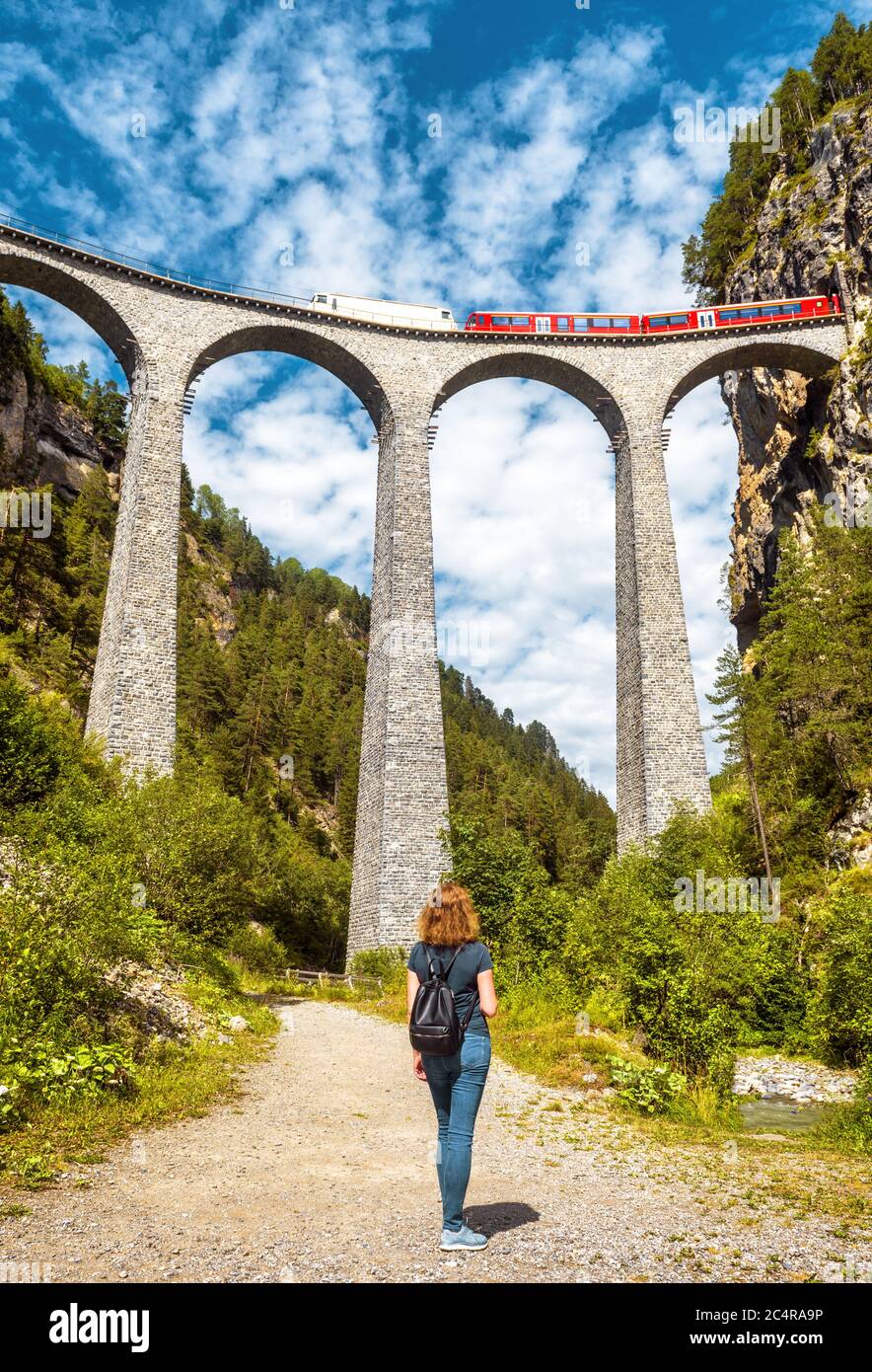 Landwasser Viadukt im Sommer, Filisur, Schweiz. Junge Frau schaut auf das berühmte Wahrzeichen der Schweizer Alpen. Rote Bahn fährt auf der hohen Eisenbahnbrücke in Alpin Stockfoto