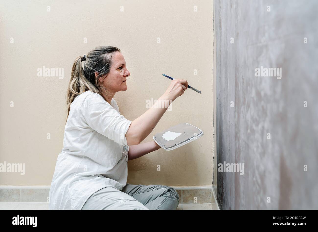 Blonde barfuß Frau sitzt auf ihrem Boden beginnt malen eine braune Wand in eine graue Farbe mit einem kleinen Pinsel. Die Frau trägt ein weißes Hemd und ein Stockfoto