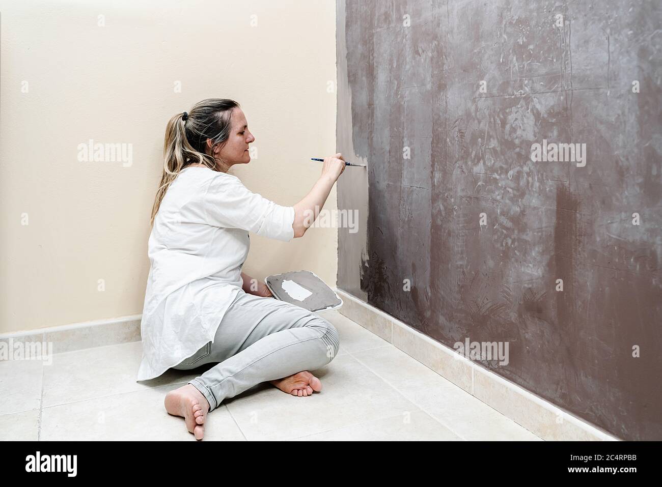 Blonde barfuß Frau sitzt auf ihrem Boden beginnt malen eine braune Wand in eine graue Farbe mit einem kleinen Pinsel. Die Frau trägt ein weißes Hemd und ein Stockfoto