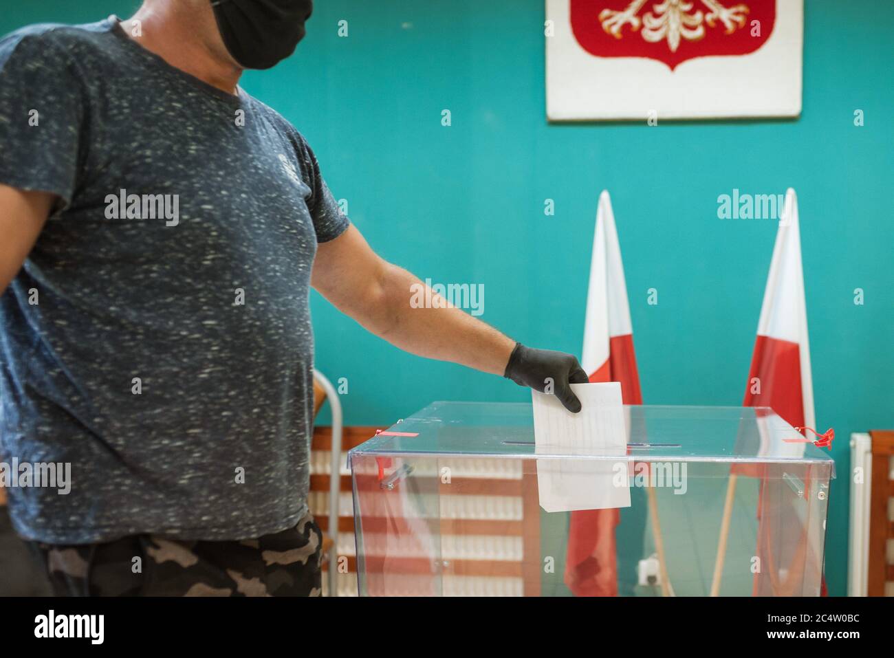 Mann mit Gesichtsmaske und Handschuhen, wegen Pandemie Covid-19, warf eine Karte mit einer Stimme an die Wahlurne während der Wahlen. Im Hintergrund polieren embl Stockfoto